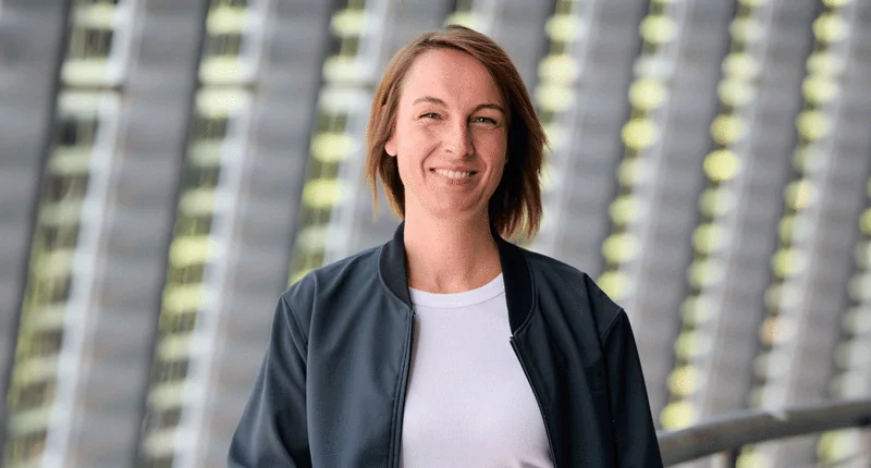 Anne-Kathrin Laufmann lächelt selbstbewusst vor einer modernen, lichtdurchfluteten Architektur und steht für Vielfalt im Fußball.
