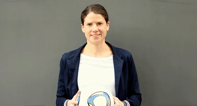Annike Krahn steht lächelnd mit einem Fußball in der Hand vor einem neutralen Hintergrund und verkörpert den Frauenfußball.