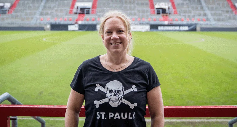 Sandra Schwedler steht lächelnd auf dem Fußballfeld, während sie ein T-Shirt mit Piratendesign trägt und den Sport feiert.